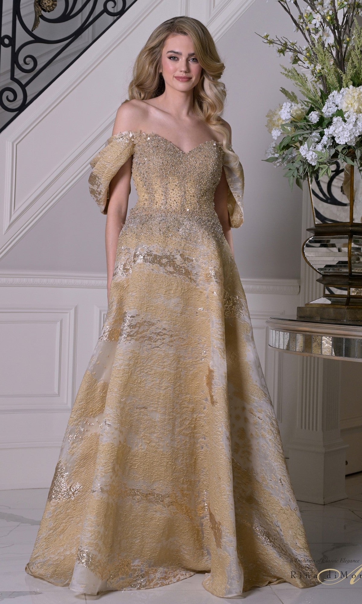 A woman stands indoors by a staircase and floral arrangement, wearing the Rina Di Montella RD3405—a gold and cream off-the-shoulder organza formal gown with an A-line skirt and subtly beaded bodice. Her wavy hair frames her face.